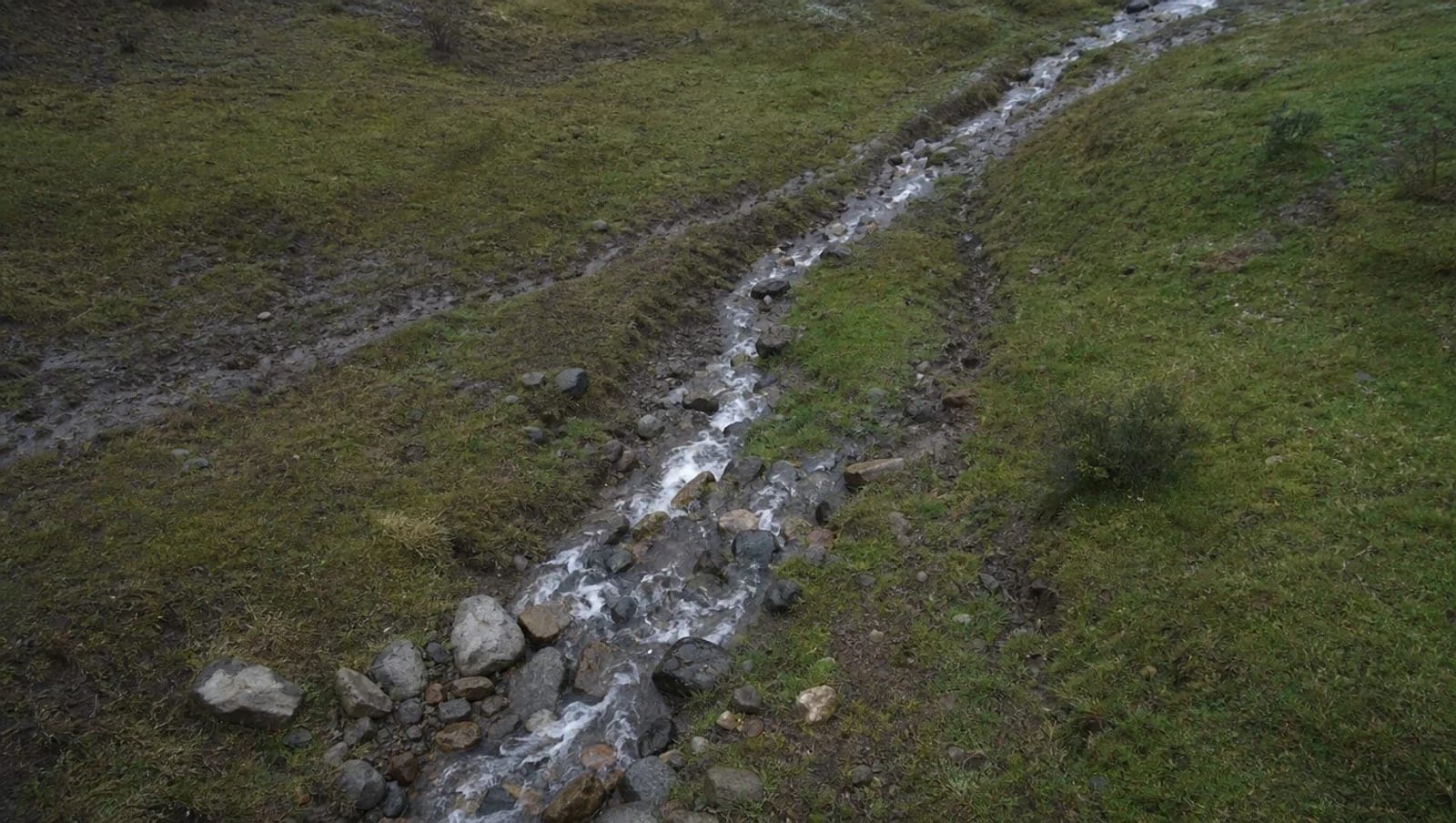 Escurrimiento de agua entre piedras atravesando un terreno con pasto, con la huella del cauce marcada en el suelo